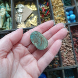 Hand holding a green scarab stone with carvings against a background of assorted beads and jewelry.