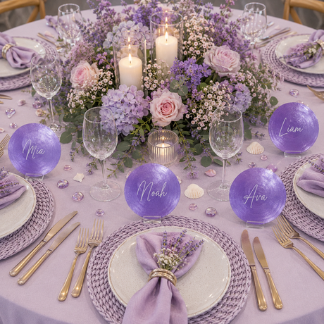 A beautifully styled lavender and purple table setting featuring round purple shell place cards with names like Mia, Noah, Ava, and Liam, decorated with purple hydrangeas, pink roses, white candles, and gold flatware on woven placemats.