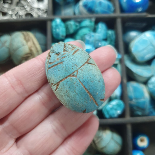 Turquoise scarab beetle bead stone held in a hand with a display of similar stones in the background