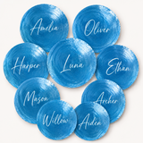 A set of round blue watercolor-style place cards with white script names including Amelia, Oliver, Harper, Luna, Ethan, Mason, Willow, Archer, Aiden, and Will, arranged on a light cream background.