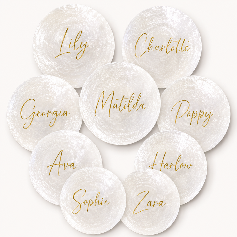 Nine round white textured shells with gold script names printed on them, arranged in a circular pattern on a light cream background. The names include Lily, Charlotte, Georgia, Matilda, Poppy, Ava, Harlow, Sophie, and Zara.