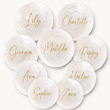 Nine round white textured shells with gold script names printed on them, arranged in a circular pattern on a light cream background. The names include Lily, Charlotte, Georgia, Matilda, Poppy, Ava, Harlow, Sophie, and Zara.