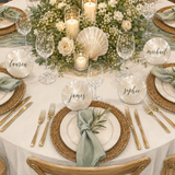 A beautifully styled wedding table setting with round white shell name cards displaying names like lauren, michael, james, and sophie, arranged on a white tablecloth with woven placemats, gold flatware, green napkins, and floral centerpieces.