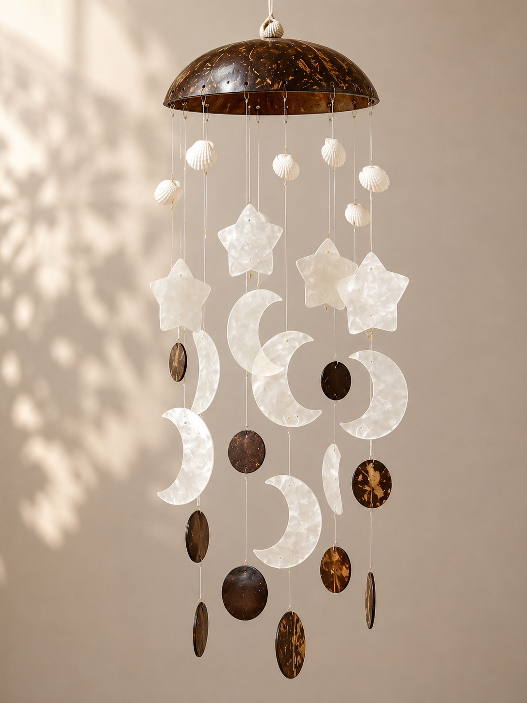A hanging celestial mobile with a dark brown coconut shell top and moon and star shapes in white and brown, suspended against a beige wall with soft leaf shadows.