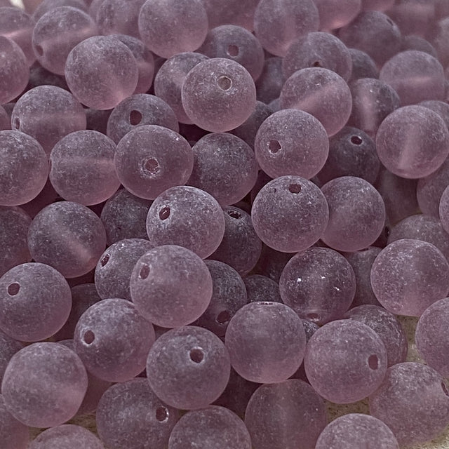 A close-up pile of small, round, frosted glass beads in a dusty lavender-purple color, each featuring a pre-drilled hole.