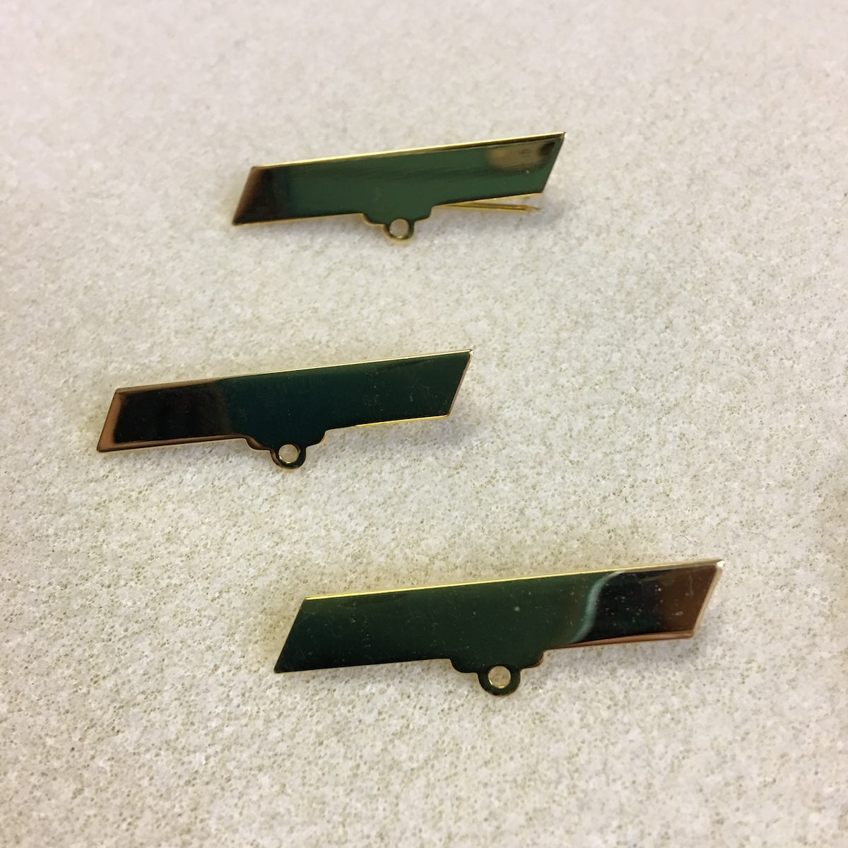 Three green and gold rectangular metal brooch fittings on a textured surface