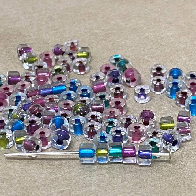 Assorted colorful beads on a beige surface including kiwi berry blue purple teal with some on a pin to show shae