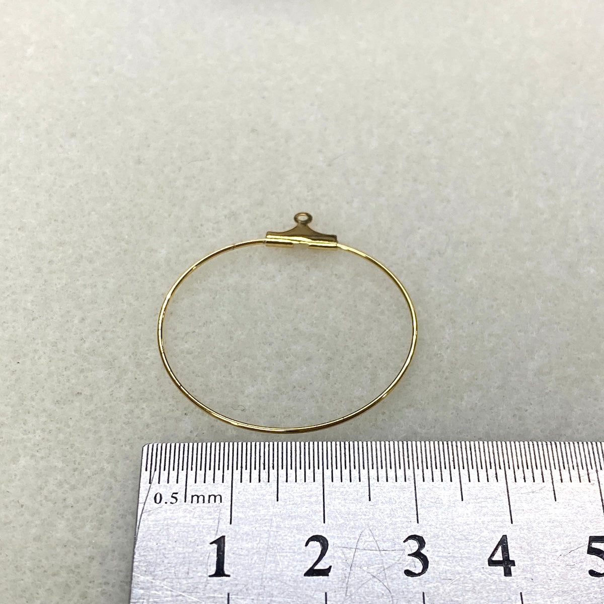 Gold circular metal frame with a small hook on top, next to a ruler for scale on a light gray background.