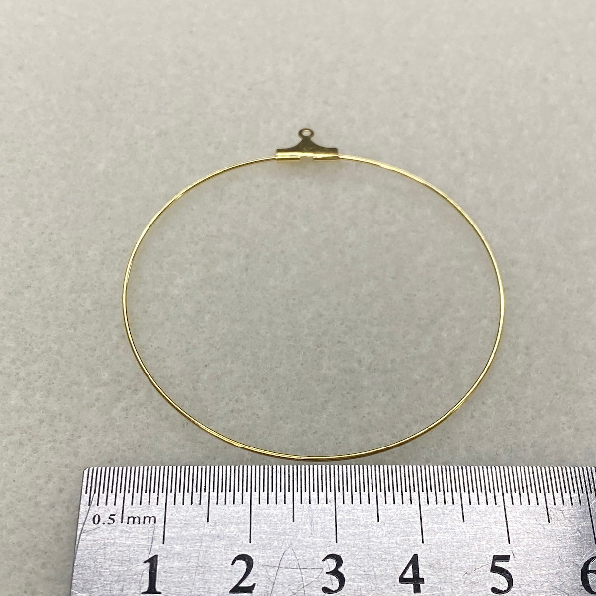 Gold circular ear hoop with a hook on a gray background, measured against a ruler.