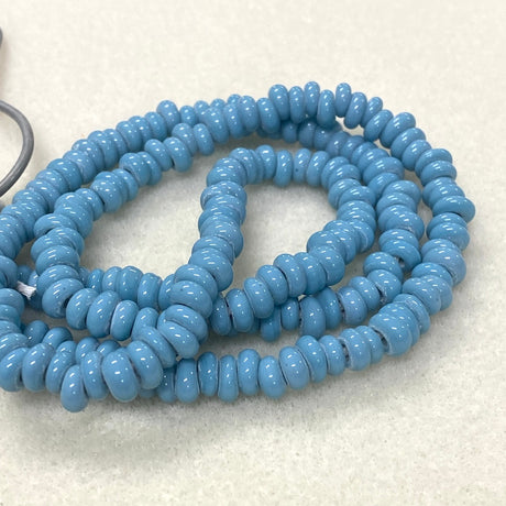 A coiled strand of glossy, disc-shaped turquoise beads in a light blue color, displayed on a textured off-white surface.