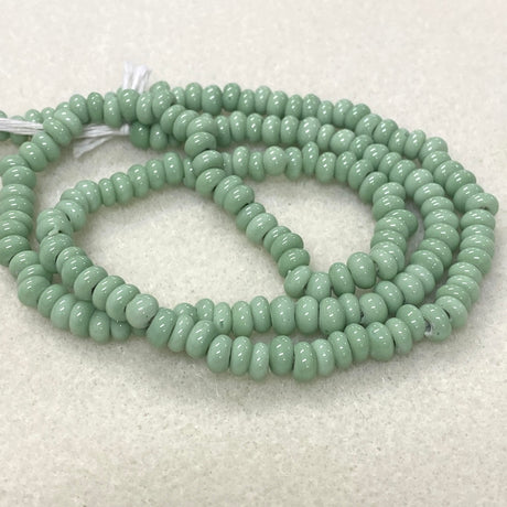 A coiled strand of small, glossy, light green rondelle donut shape  beads on a textured off-white background.