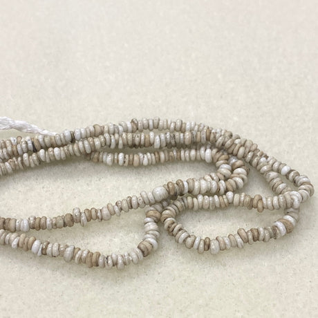 A strand of small, irregularly shaped beads in natural beige, cream, and off-white tones, arranged in loops on a textured light beige background.