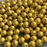 A close-up pile of small metallic gold round beads with holes, filling the frame against a dark textured background.