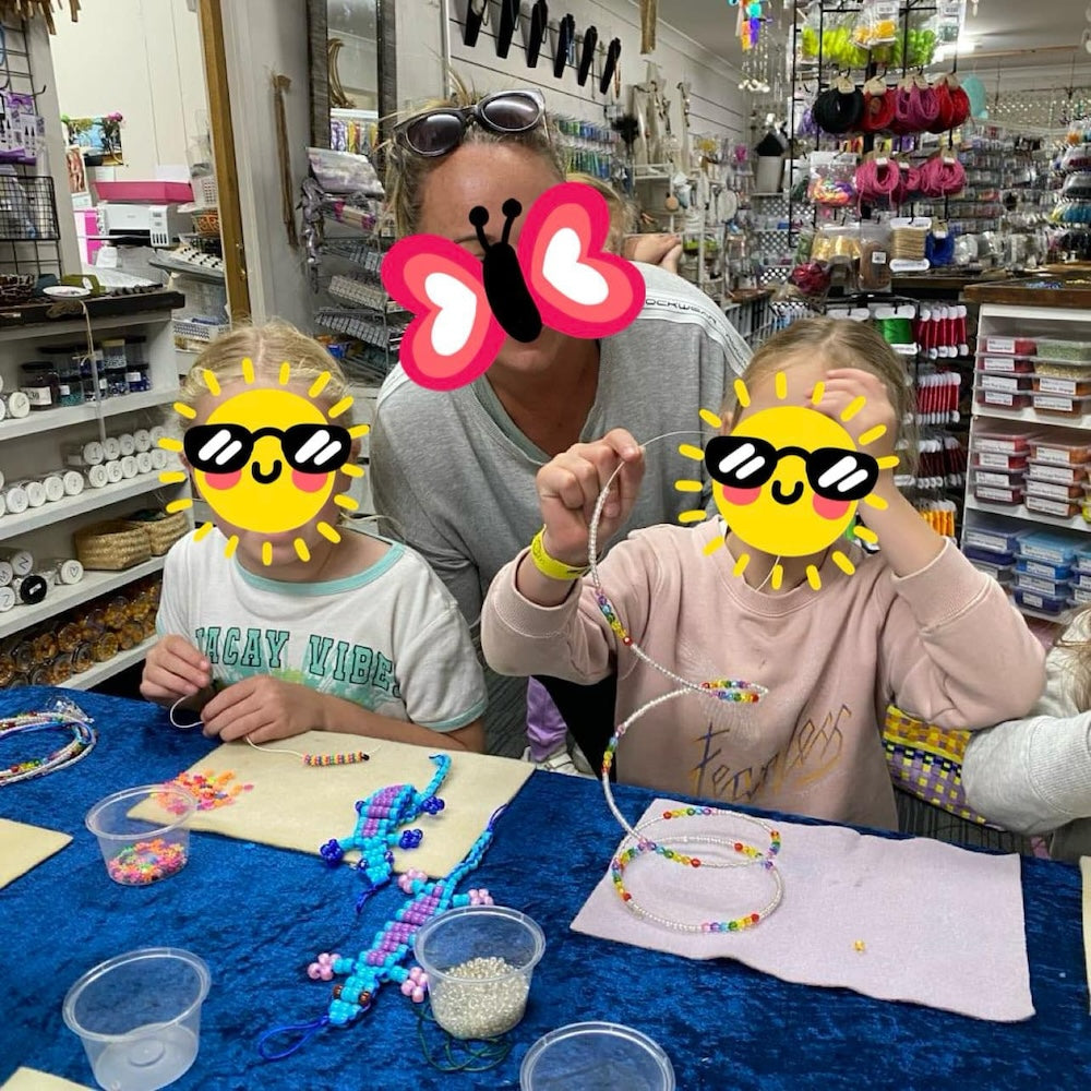 Children and mother at the work table making animals, hanging decorations and jewelry at a workshop run by Bead Shack showing lots of stock in the background of the shop.  Faces are covered with comic icons