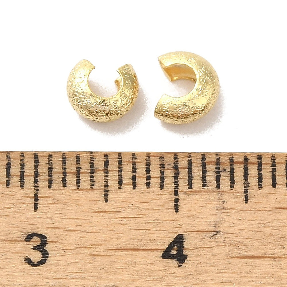 close up of 2 crimp covers with textured gold surface next to a wood ruler with sizes