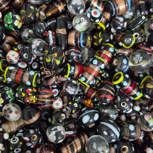 A dense pile of handmade glass lampwork beads featuring various patterns including stripes, polka dots, swirls, and floral motifs in black, brown, red, white, green, yellow, and blue.