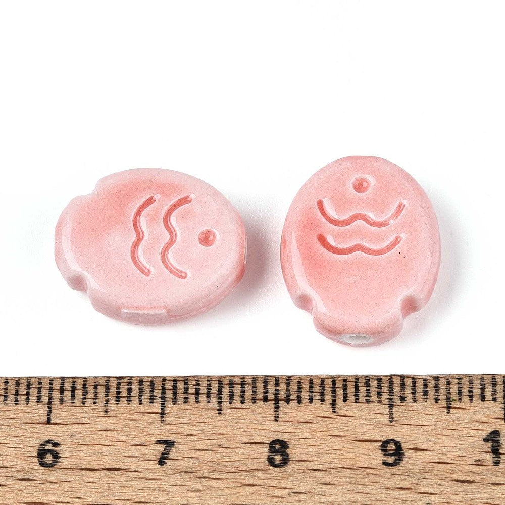 Two pink ceramic fish beads with wave designs on a white background, measured against a wooden ruler.