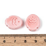 Two pink ceramic fish beads with wave designs on a white background, measured against a wooden ruler.