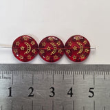 Three red beads with gold patterns on a metal ruler for scale.
