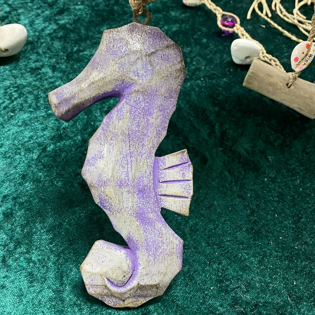 A hand painted and hand carved wooden seahorse decoration in purple color with shabby chic painting, displayed on a green background with other decorative items.